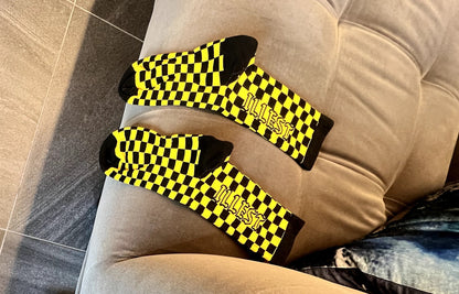 Two pairs of shoes and three socks in our black and yellow checkered style on a couch and white Hoka sneakers on the floor.