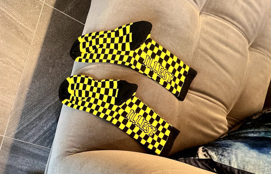 Two pairs of shoes and three socks in our black and yellow checkered style on a couch and white Hoka sneakers on the floor.